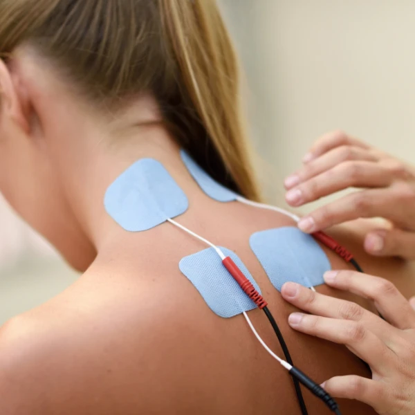 Nahaufnahme einer Frau von hinten, auf deren Nacken und oberen Rücken mehrere blaue Reizstromeletroden für eine neurologische Untersuchung angebracht werden. Eine Person platziert die Elektroden vorsichtig per Hand.